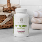 60 count capsule bottle of Gut Restore with Betaine and HCL sitting on a dark wooden countertop with green house plants and white tile background.