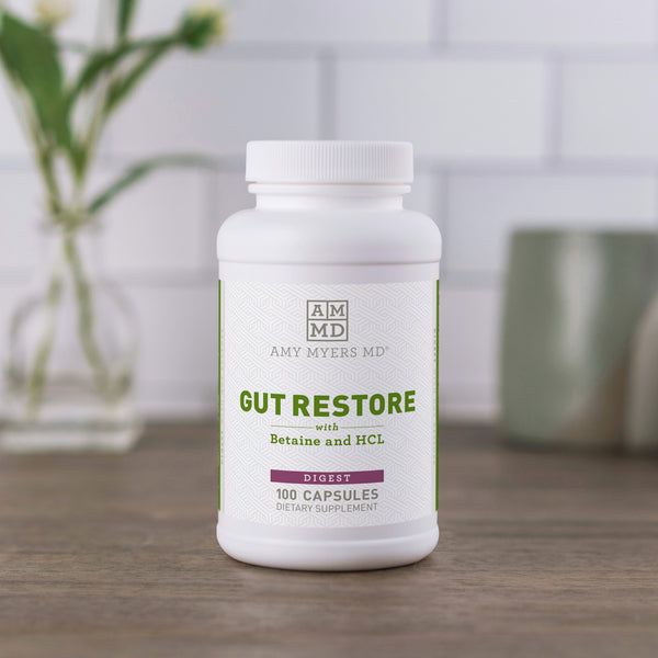 100 count capsule bottle of Gut Restore with Betaine and HCL sitting on a dark wooden countertop with green house plants and white tile background.