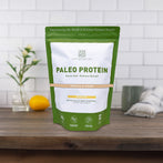 Resealable bag of AMMD Vanilla Bean flavored Paleo Protein sitting on a wooden countertop, with white tile background.
